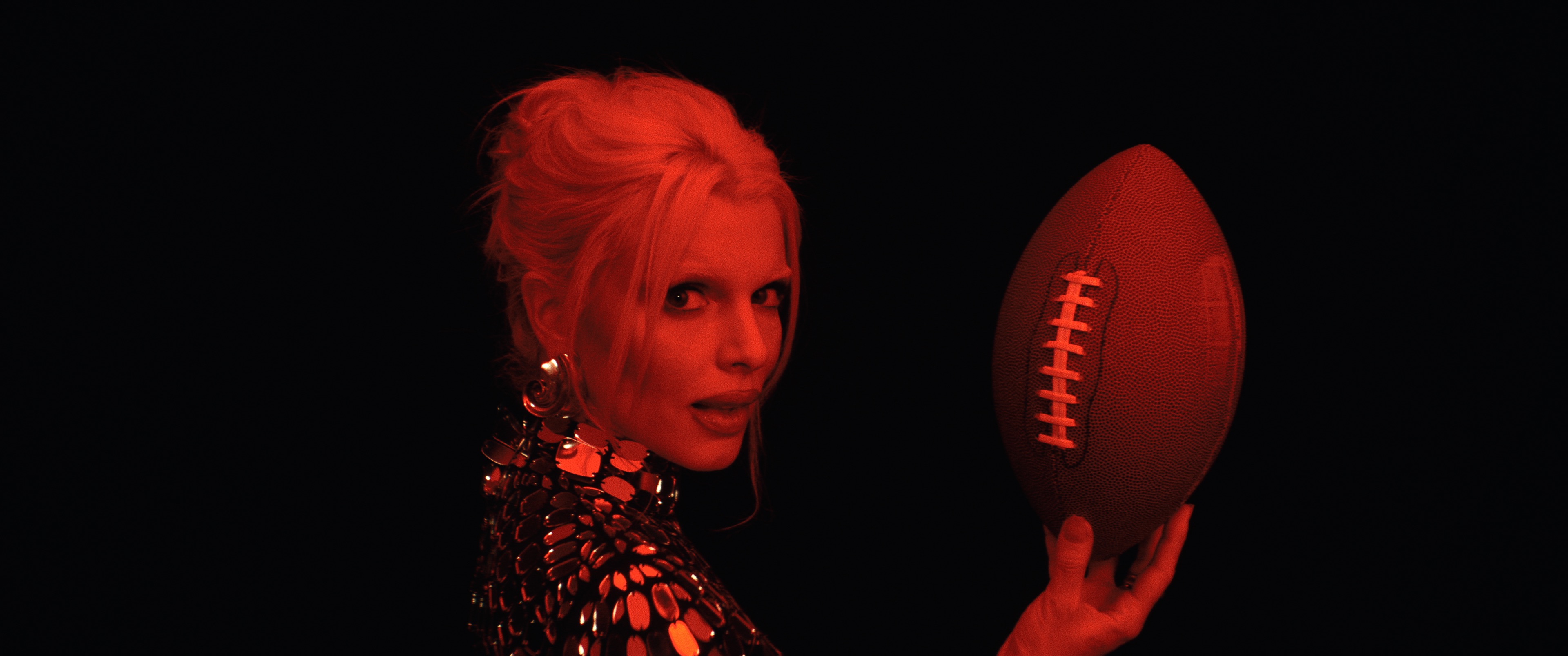 Actor Julia Fox in character cast in red light holding a foot ball.