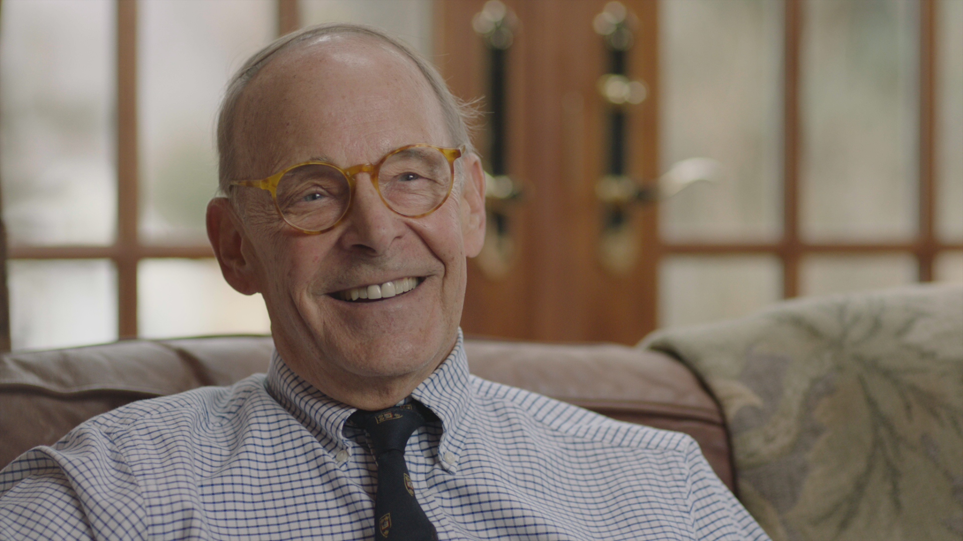 Dr. James Sehn in the 2019 documentary series LORENA sitting on a couch bespectacled, and wearing a collared shirt and tie.