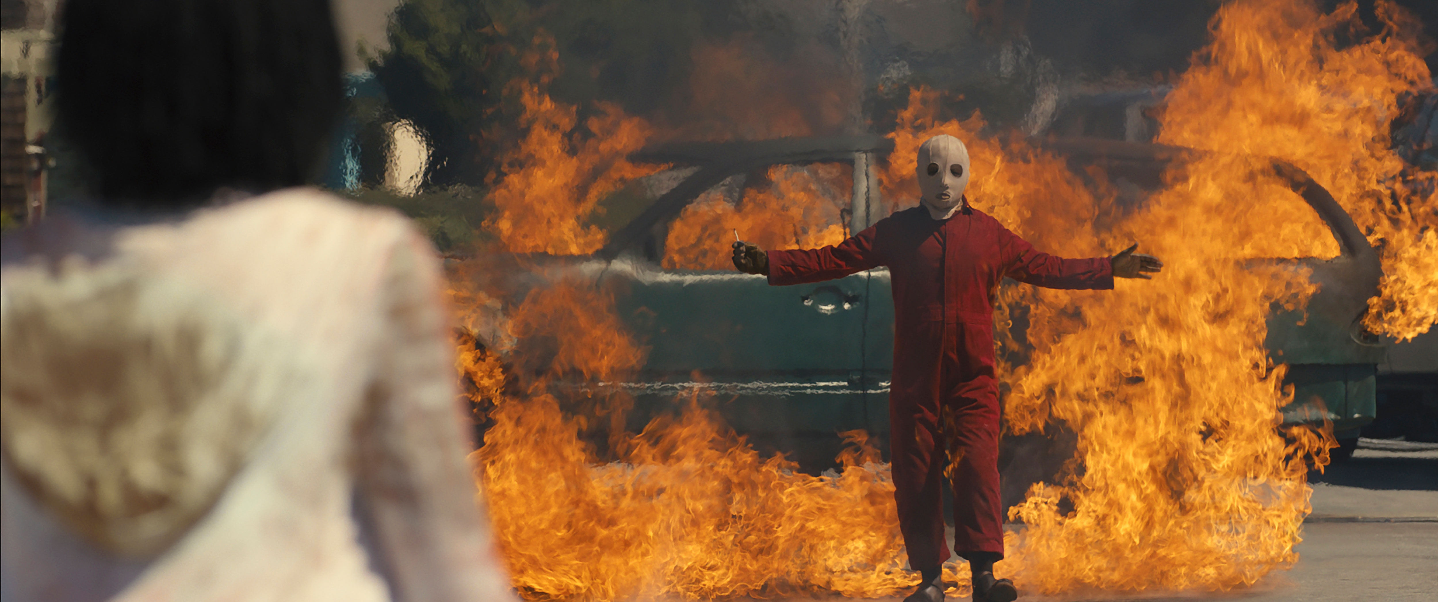 Evan Alex in the 2019 film US standing, arms extended, in front of a burning car, and wearing a red jumpsuit and white mask.