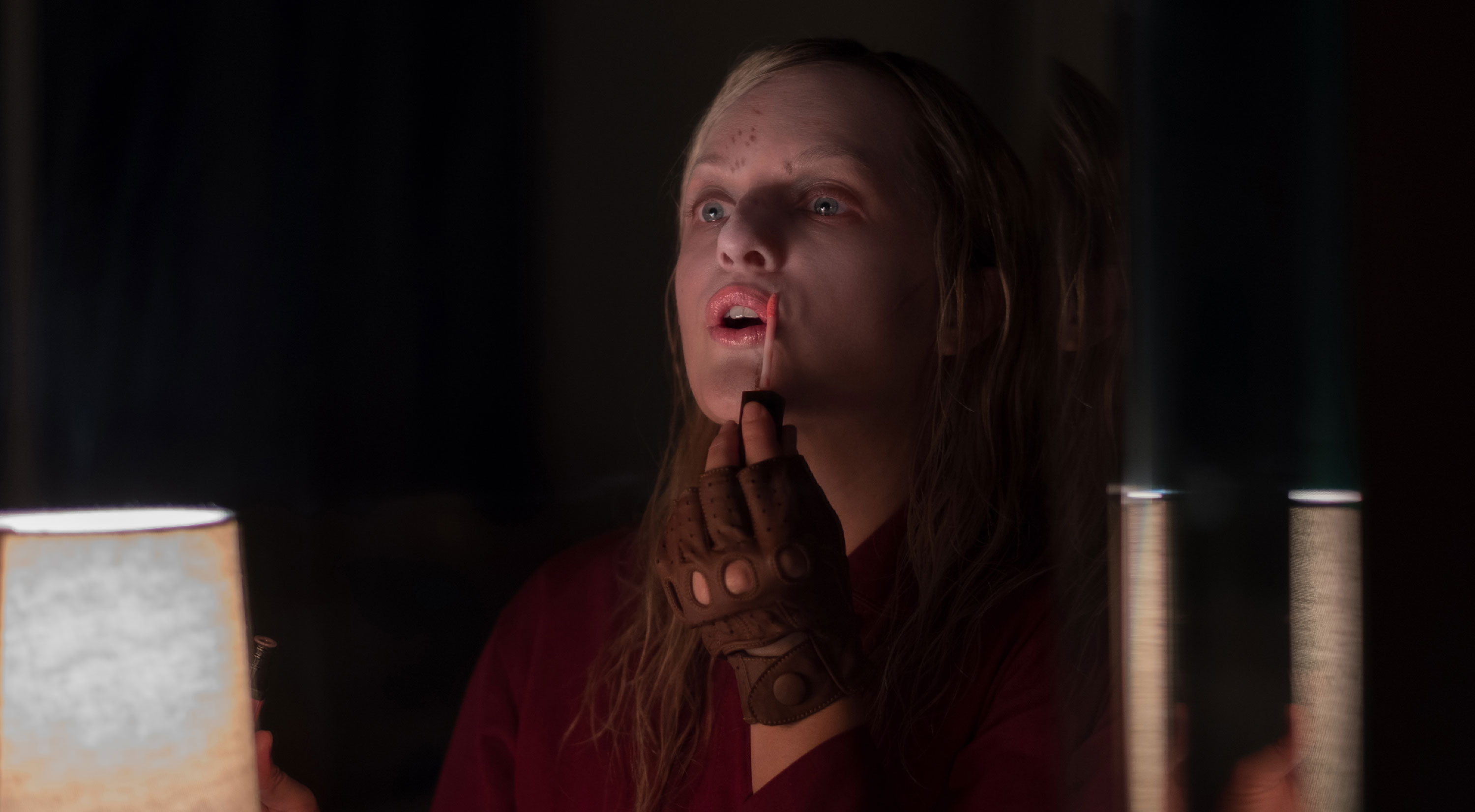 Elisabeth Moss in the 2019 film US wearing red jumpsuit and leather driving glove and applying lipgloss at a mirror.