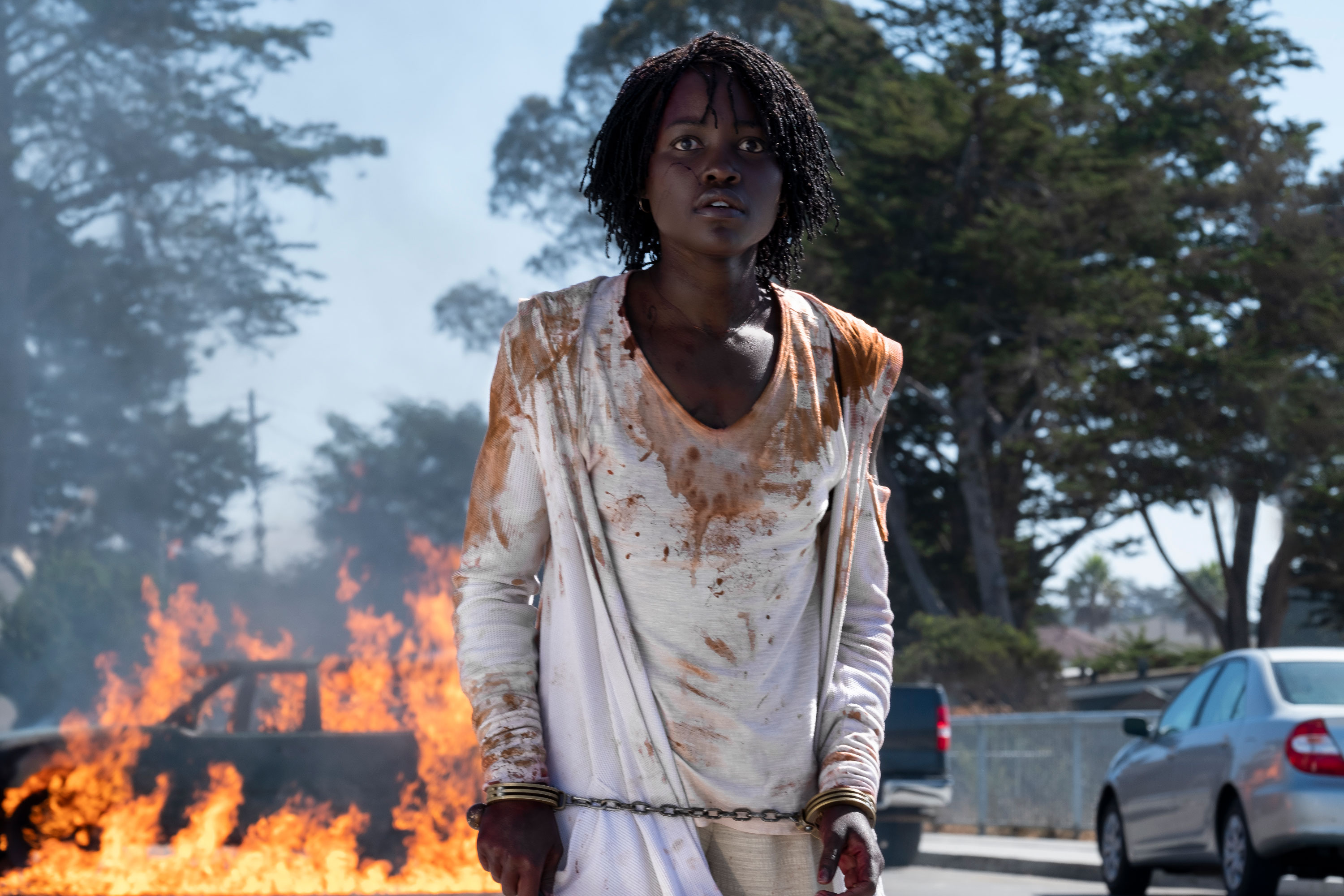 Lupita Nyongo'o in the 2019 film US standing in front of a burning car wearing stained clothes and handcuffs.