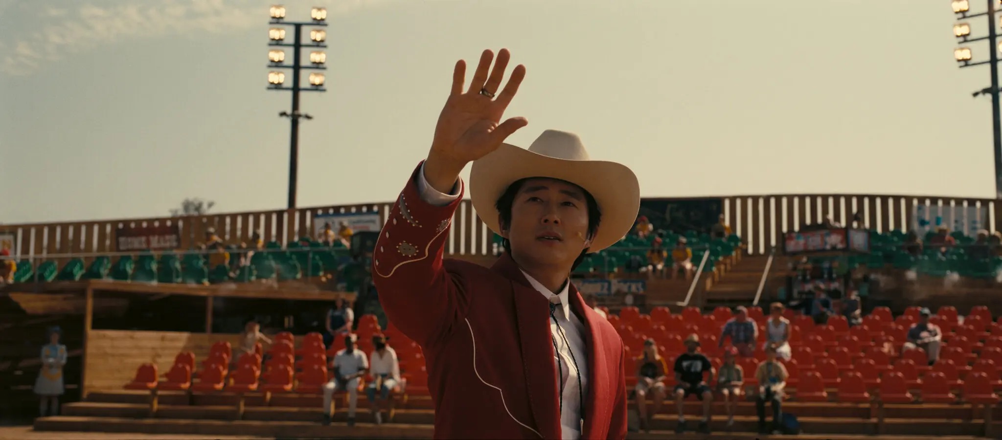 Steven Yeun in the middle of a huge stadium looking directly at the sky with his hand up.