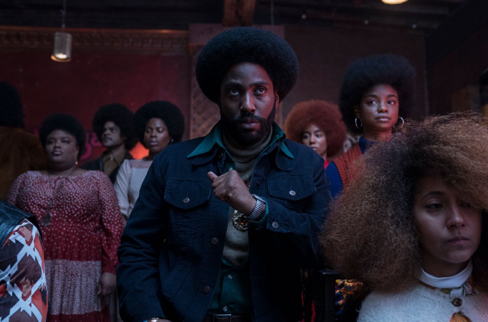 John David Washington in the 2018 film BLACKKKLANSMAN leaning on table in crowd of people.