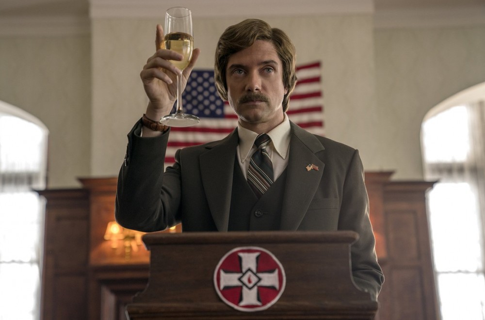 Topher Grace in the 2018 film BLACKKKLANSMAN standing at a podium and lifting a glass of champagne.